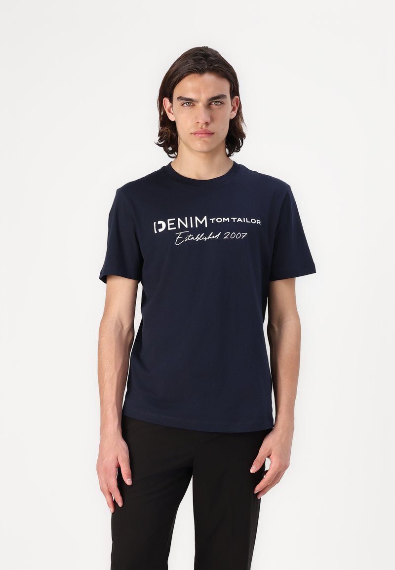 Jūras zila T-krekls, izgatavots no kokvilnas, ar īsām piedurknēm un apaļu kakla izgriezumu. Priekšpusē ir uzraksti "DENIM TOM TAILOR" un "Izveidots 2007."