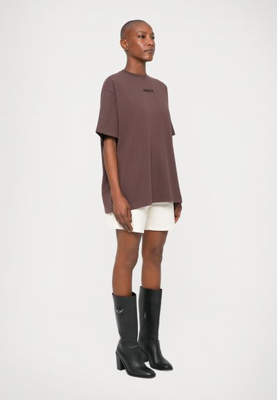 Camiseta de algodón marrón de gran tamaño con mangas cortas, que presenta un pequeño logo en el pecho, combinada con shorts blancos y botas negras hasta la rodilla.