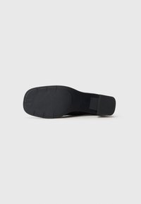 Suola di una scarpa nera con tacco quadrato, che mostra una superficie in gomma testurizzata e leggere scanalature per la trazione.