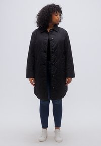 Μαύρο μακρύ παλτό με καπιτονέ ύφασμα, γιακά, κλείσιμο με κουμπιά μπροστά και δύο πλαϊνές τσέπες, φορεμένο πάνω από μαύρο τοπ και μπλε τζιν.