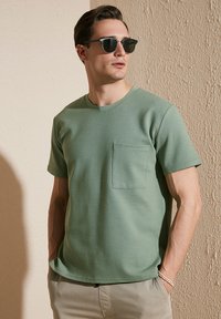 T-shirt verde con texture, maniche corte e taschino sul petto, abbinato a pantaloni chiari e occhiali da sole scuri.