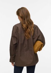 Brązowa, oversize koszula w paski z długimi rękawami i plisą z tyłu, zestawiona ze skórzaną, jasnobrązową torebką trzymaną pod pachą. Widoczne dżinsy.