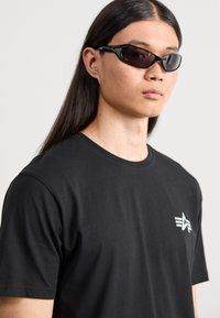 Schwarzes Baumwoll-T-Shirt mit rundem Halsausschnitt, ausgestattet mit einem kleinen weißen Logo auf der Brust. Das Modell trägt elegante, kantige schwarze Sonnenbrillen.