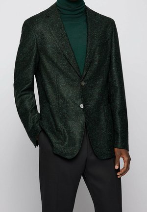 Mężczyzna w ciemnozielonym, teksturowanym blezerze, do którego dopasowany jest zielony golf, oraz czarnych spodniach, z jedną ręką w kieszeni, na jednolitym tle.