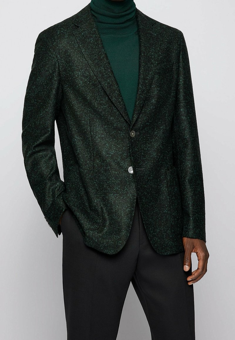 Homme portant un blazer texturé vert foncé sur un col roulé vert assorti et un pantalon noir, une main dans la poche, sur un fond uni.