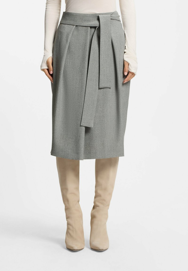 Jupe plissée grise avec un détail de taille à nouer, en tissu texturé. Portée avec de hautes bottes beiges, mettant en valeur la longueur de la jupe.