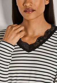 Gestreiftes Langarmshirt in Creme und Schwarz mit einem Spitzenkragen im V-Ausschnitt und gewellten Kanten. Glatte Textur und figurbetonter Schnitt.