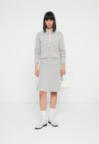 Cardigan accorciato grigio chiaro e gonna al ginocchio realizzati in un morbido e peloso tessuto; abbinati a calzini e scarpe bianche, mentre tiene una piccola borsa bianca.