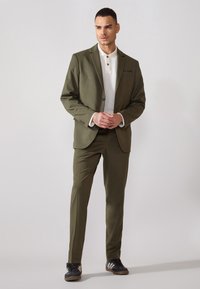 Olivgrüner Anzug mit taillierter Passform, ausgestattet mit einem Ein-Knopf-Verschluss, kombiniert mit einem weißen Polohemd und schwarzen Sneakers mit weißen Streifen.