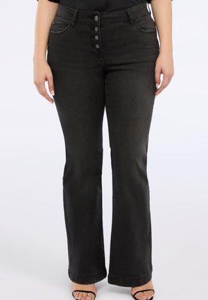 Vrouw in hoge zwarte jeans met vier knopen aan de voorkant, zijzakken en zwarte hoge hakken op een witte achtergrond.