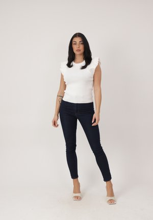 Kobieta o długich ciemnych włosach stoi, nosząc białą bezrękawnikową bluzkę z falbanami, ciemne obcisłe dżinsy i beżowe sandały na obcasie z odkrytymi palcami na jednolitym tle.