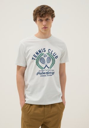 Jonge man met een wit T-shirt met tennisclub afbeelding en een bruine broek, staand met handen in de zakken tegen een effen achtergrond.