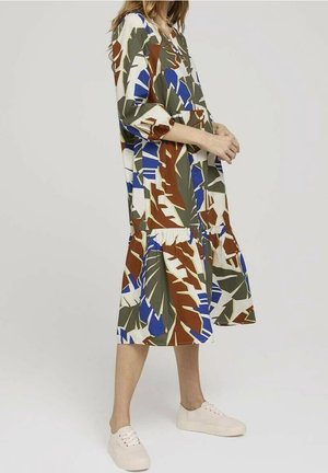 Vrouw draagt een losse, knielange jurk met blauwe, bruine, groene en witte abstracte bladpatronen en witte sneakers op een egale achtergrond.