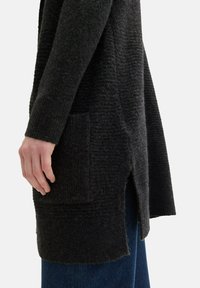 Svart stickad cardigan med ett texturerat mönster, sidoflikar och framfickor. Tyget verkar mjukt och varmt.