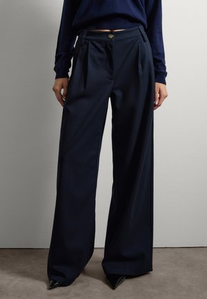 Marinefarbene weite Hosen aus glattem Stoff, mit Vorderfalten, einem Knopfverschluss und Seitentaschen. Scharfe, spitze schwarze Stöckelschuhe vervollständigen das Outfit.