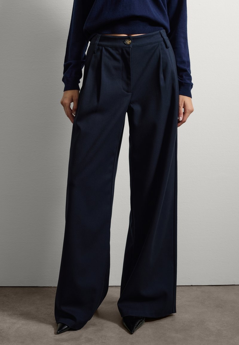 Pantalons large en tissu lisse de couleur marine, avec des plis avant, une fermeture à bouton et des poches latérales. Des talons hauts noirs pointus complètent le look.