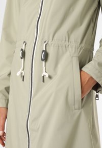 Jasnozielona wodoodporna kurtka z przednim zamkiem błyskawicznym, ściągaczem w talii i bocznymi kieszeniami z czarnymi akcentami metalowymi. Gładka tekstura.