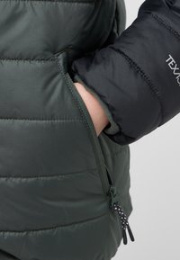 Giacca a vento verde con una texture liscia, dotata di una tasca con zip e maniche nere. La zip della tasca ha un tirante decorativo.