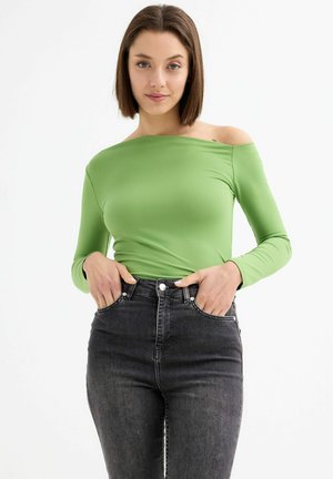 Top verde de manga larga y sin hombros, hecho de una tela elástica, combinado con jeans de mezclilla oscuros de tiro alto, presenta un diseño ajustado y una textura suave.