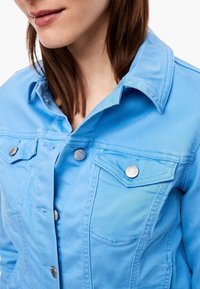 Hellblaues Denim-Jackett mit einem Kragen, silbernen Druckknöpfen und zwei aufgesetzten Brusttaschen. Glatte Textur über die gesamte Oberfläche.