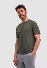 T-shirt verde oliva a maniche corte con scollatura rotonda; realizzato in cotone morbido. Abbinato a pantaloni della tuta grigio chiaro con tasche laterali.