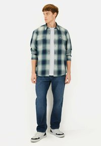 Gekraagde shirt in een groen en marineblauw ruitpatroon, met een knoopsluiting aan de voorkant en twee borstzakken, gedragen over een wit t-shirt en blauwe jeans.