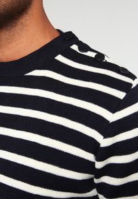 Sweter dzianinowy w niebiesko-białe paski z okrągłym dekoltem, teksturowanym materiałem i czarnymi guzikami na ramieniu.