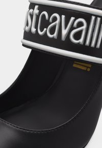 Μαύρο ψηλοτάκουνο παπούτσι με φαρδιά λουρίδα που φέρει ανάγλυφα λευκά γράμματα «Just Cavalli» στην επιφάνεια.