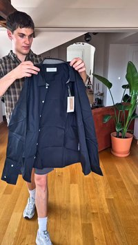 Schwarzes Hemd mit Knopfleiste, strukturiertem Kragen, langen Ärmeln und einem Etikett an der Seite. Der Stoff wirkt glatt und leicht.