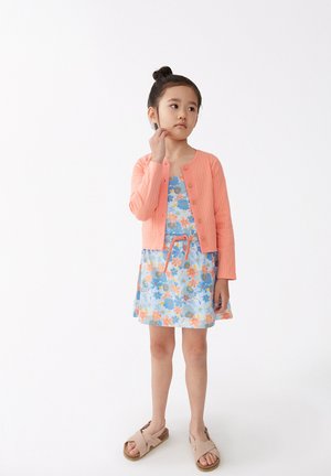 Cárdigan de coral con canalé, vestido ligero azul con estampado floral y acentos en naranja, combinado con sandalias beige, estilizado con un peinado de moño simple.