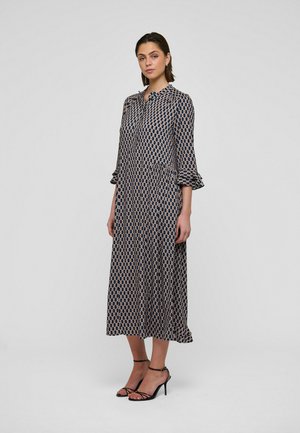 Maxikleidis mereva sügavsinises toonis, kreemika geomeetrilise mustriga, pikkade varrukatega, volangikujuliste mansettidega, nööpidega esiküljel ja lehviva seelikuga. Kandes mustade kontsakingadega.