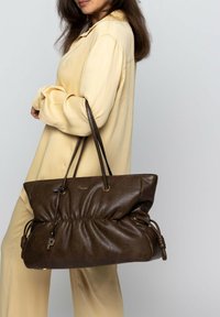 Bruine leren tote bag met een zachte, gekreukte textuur, trekkoorddetail en een decoratieve sleutelhangeraccent. Grote maat met twee schouderbanden.