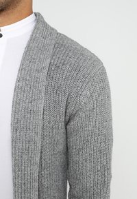 Cardigan grigio lavorato a maglia con una texture morbida, apertura frontale e dettagli a costine sulle maniche. Presenta una silhouette rilassata e un design informale.