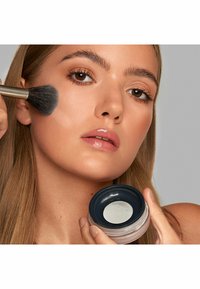 Translucentes Puder in einem runden Behälter mit einem dunklen Deckel. Ein Pinsel trägt das Produkt auf die Wange auf und hebt die glatte Hautstruktur hervor.