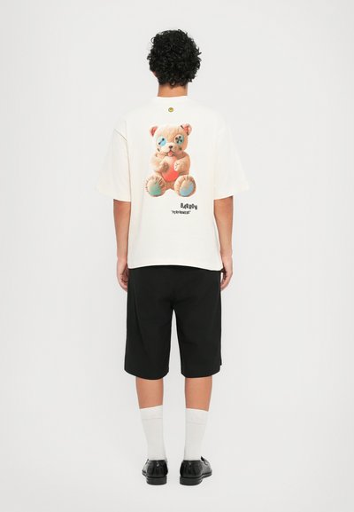 Camiseta de algodón en beige claro con un gráfico de oso de peluche en la parte posterior, combinada con pantalones cortos negros y calcetines blancos. Diseño simple, corte relajado.