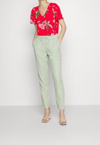 Top floreale a V in rosso con fiori rosa e verdi, abbinato a pantaloni leggeri verdi chiaro con vita elastica e cordoncino. Sandali con tacco neutri.