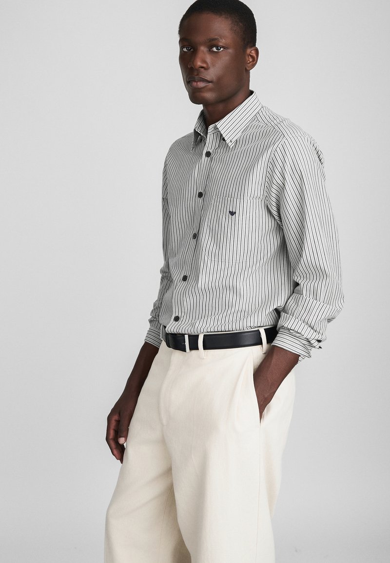 Stripet skjorte med knapper i svart og hvitt, med en brystlomme og svarte knapper. Kombinert med kremfargede bukser og et svart belte.