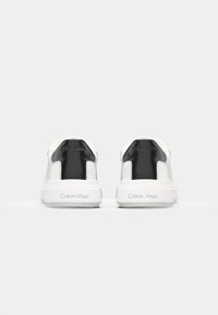 White sneakers with a black back panel, smooth leather material, flat sole, and embossed "Calvin Klein" branding on the heel.
