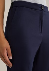 Primo piano della mano di una persona che riposa accanto a pantaloni su misura blu navy con una vita dotata di bottone e tasca laterale.