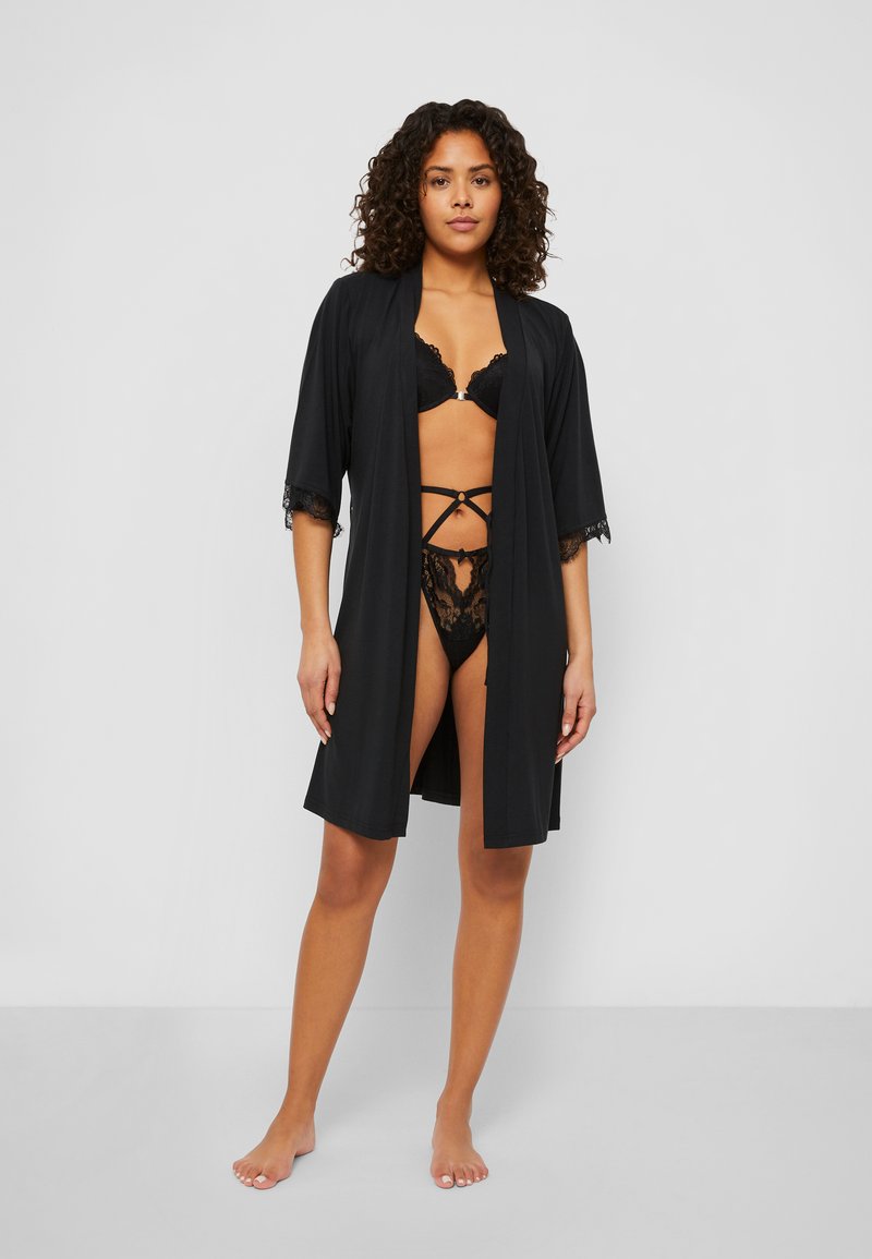 Kimono en satin noir avec des finitions en dentelle et des manches courtes, porté sur un bralette en dentelle noire et un string, présentant un design superposé.