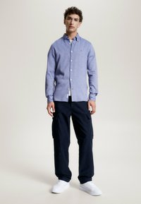 Camicia a maniche lunghe azzurro chiaro con colletto button-down, abbinata a pantaloni cargo scuri. L'outfit è completato da sneaker bianche.