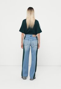 Una persona con lunghi capelli biondi sta in piedi di spalle, indossando una maglietta verde scuro a maniche corte, jeans blu e verdi a blocchi di colore e scarpe grigie.