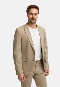 Beige Anzugjacke aus glattem Stoff, mit Reverskragen, zwei Knöpfen und seitlichen Taschen; getragen über einem weißen T-Shirt.