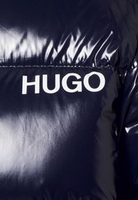 Marineblå pufferjakke med en blank finish, der har et fremtrædende hvidt "HUGO" logo og syninger på brystområdet.