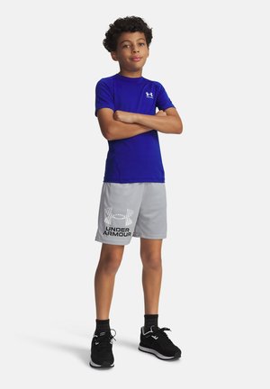 Ragazzo in piedi con le braccia incrociate indossa una maglietta blu Under Armour, pantaloncini grigi con logo, calzini neri e scarpe da ginnastica nere su sfondo bianco.