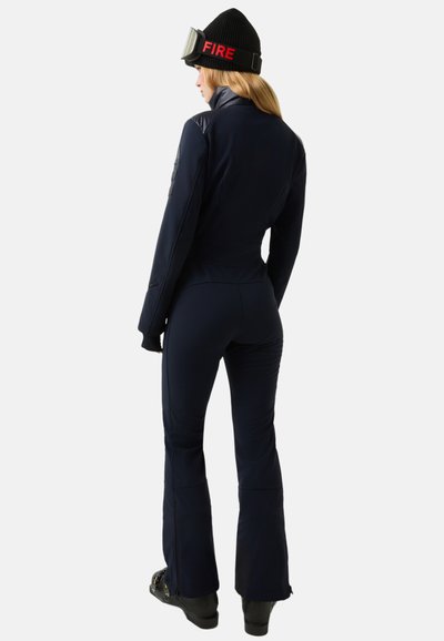 Tmavě modrý lyžařský oblek s přiléhavým střihem, s vysokým límcem, kapsami na zip a rozšířenými kalhotami. Nošený s černou čepicí s nápisem "FIRE."