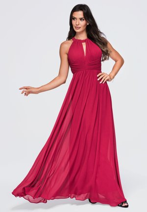 Femme portant une robe rouge longue jusqu'au sol, sans manches, avec un corsage plissé et un décolleté en forme de trou de serrure, posant avec une main sur la hanche et l'autre tendue.