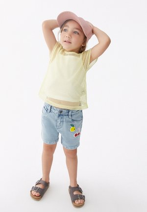 Top de malla amarillo, shorts de mezclilla azul claro con bordado de frutas y sandalias grises. La gorra es rosa, añadiendo un sutil acento al atuendo.