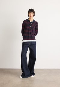 Young person stands with hands behind back wearing navy and burgundy striped long-sleeve shirt, wide-leg dark jeans, and white shoes.