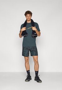 Mørk teal atletisk T-shirt med et gult logo, parret med sorte mønstrede shorts. Modellen holder et sort håndklæde om skuldrene og har sorte sneakers på.